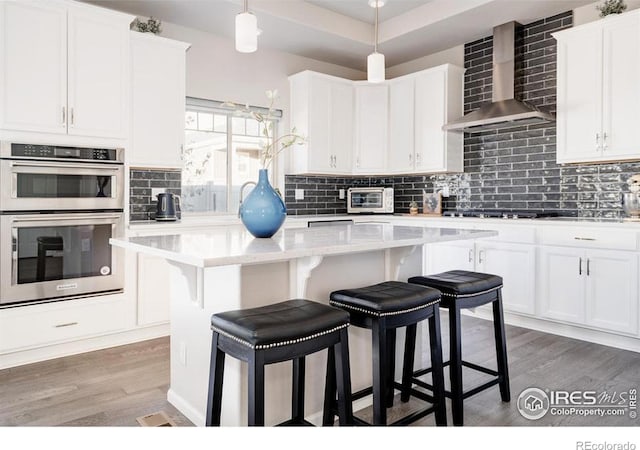 kitchen featuring stainless steel double oven, light countertops, a center island, wall chimney exhaust hood, and tasteful backsplash