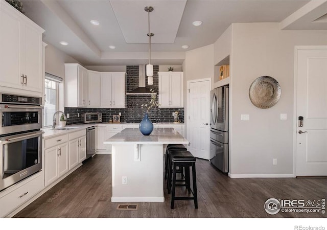 kitchen with a tray ceiling, a breakfast bar, appliances with stainless steel finishes, dark wood-type flooring, and wall chimney range hood