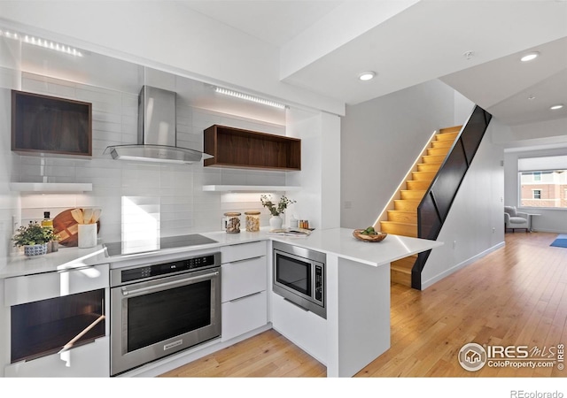 kitchen featuring kitchen peninsula, stainless steel appliances, wall chimney range hood, and light wood-type flooring