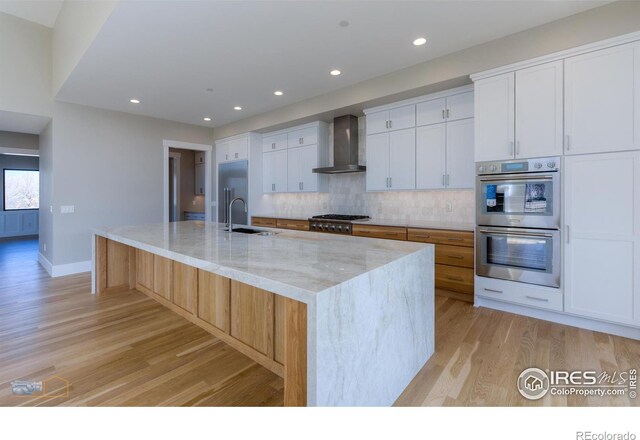 kitchen with light stone counters, stainless steel appliances, a spacious island, white cabinets, and wall chimney range hood