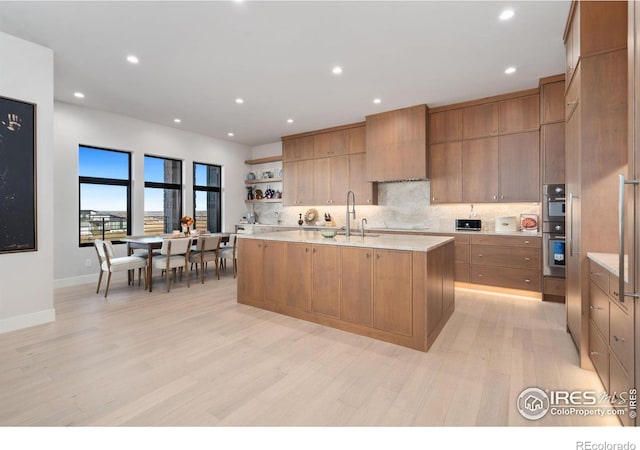 kitchen with light wood-type flooring, tasteful backsplash, custom range hood, double oven, and an island with sink