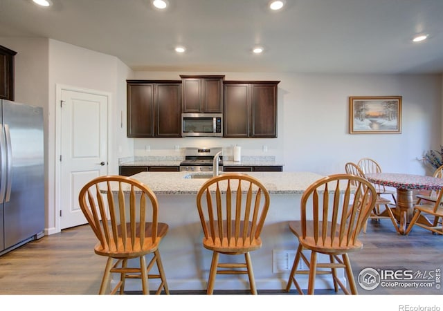 kitchen featuring a kitchen island with sink, dark hardwood / wood-style floors, light stone counters, dark brown cabinetry, and stainless steel appliances