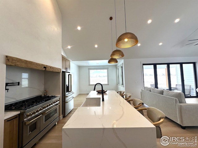 kitchen with double oven range, light hardwood / wood-style floors, a center island with sink, hanging light fixtures, and light stone counters