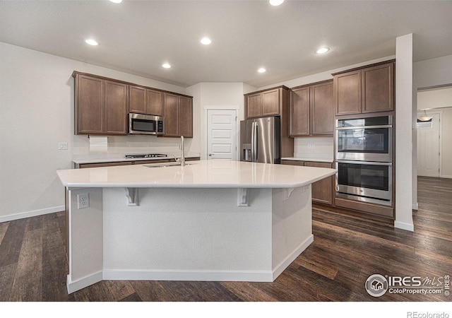 kitchen with dark brown cabinetry, sink, dark wood-type flooring, a kitchen island with sink, and appliances with stainless steel finishes