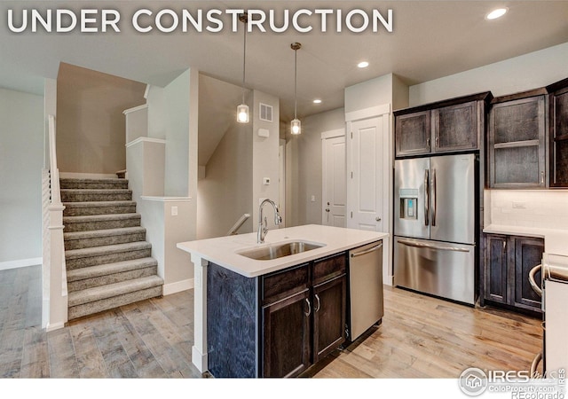 kitchen featuring light wood-type flooring, stainless steel appliances, a kitchen island with sink, sink, and hanging light fixtures