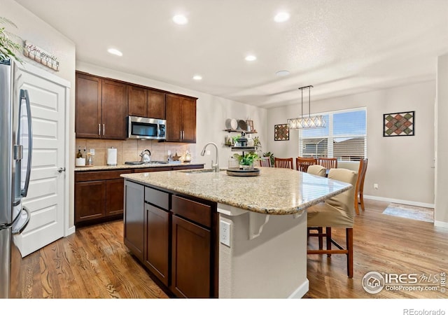 kitchen with appliances with stainless steel finishes, a kitchen island with sink, sink, decorative light fixtures, and light hardwood / wood-style flooring