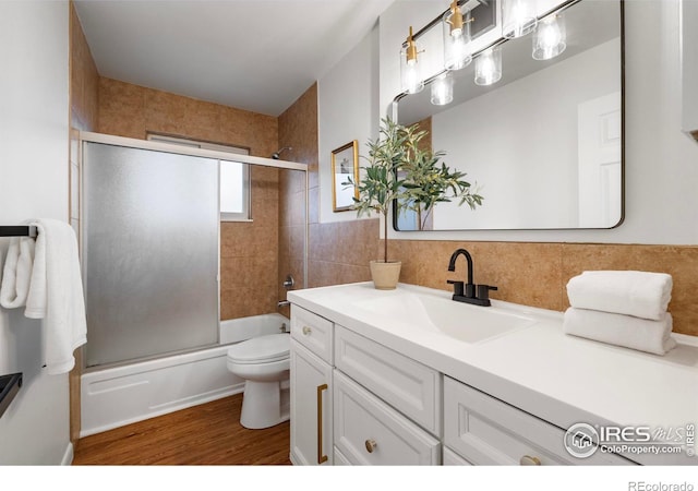 full bathroom featuring hardwood / wood-style floors, vanity, bath / shower combo with glass door, toilet, and tile walls