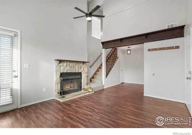 unfurnished living room featuring wood-type flooring, a towering ceiling, a stone fireplace, and ceiling fan