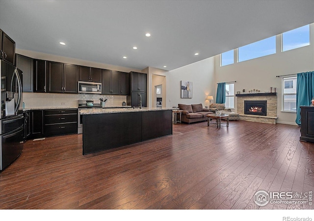 kitchen with dark hardwood / wood-style flooring, backsplash, a towering ceiling, an island with sink, and appliances with stainless steel finishes