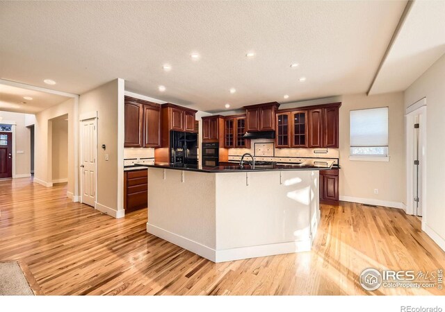 kitchen featuring a center island with sink, light wood finished floors, a sink, glass insert cabinets, and dark countertops