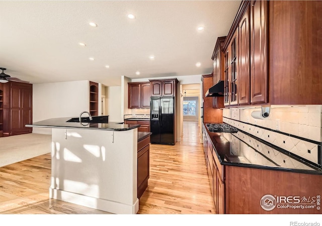 kitchen featuring a center island with sink, light wood-style floors, under cabinet range hood, dark countertops, and black fridge