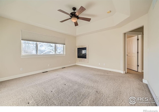 carpeted spare room with baseboards, visible vents, a tray ceiling, ceiling fan, and a glass covered fireplace