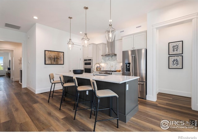 kitchen featuring a center island with sink, white cabinetry, stainless steel appliances, wall chimney exhaust hood, and hanging light fixtures