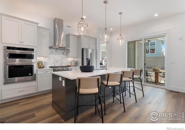 kitchen with appliances with stainless steel finishes, an island with sink, wall chimney range hood, pendant lighting, and a kitchen bar