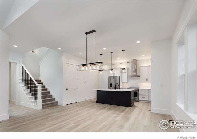 kitchen with white cabinetry, wall chimney range hood, stainless steel appliances, hanging light fixtures, and an island with sink