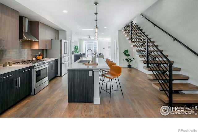 kitchen featuring sink, wall chimney range hood, dark hardwood / wood-style floors, a center island with sink, and appliances with stainless steel finishes