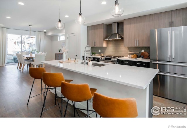 kitchen featuring sink, wall chimney exhaust hood, stainless steel appliances, dark hardwood / wood-style flooring, and a center island with sink