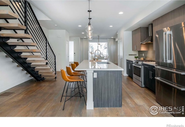 kitchen with wall chimney range hood, sink, light hardwood / wood-style flooring, an island with sink, and appliances with stainless steel finishes