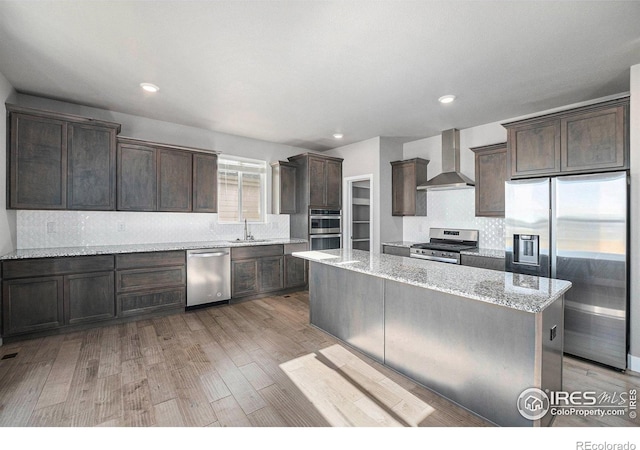 kitchen with wall chimney exhaust hood, stainless steel appliances, light stone counters, light hardwood / wood-style floors, and a kitchen island