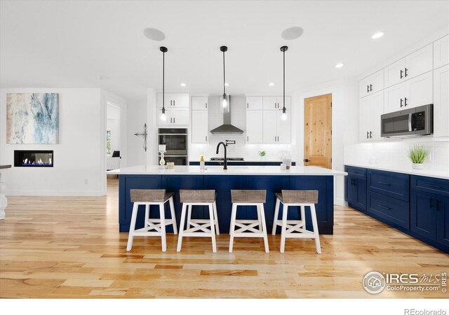 kitchen featuring white cabinets, appliances with stainless steel finishes, a center island with sink, and wall chimney range hood