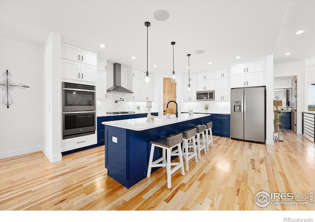 kitchen with appliances with stainless steel finishes, a kitchen breakfast bar, a kitchen island with sink, wall chimney range hood, and white cabinetry