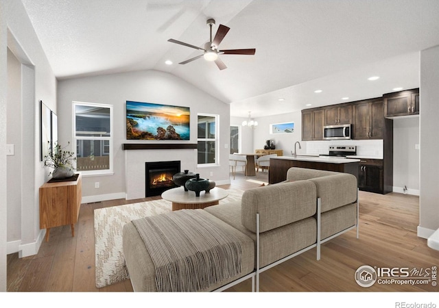 living room with light wood finished floors, lofted ceiling, a warm lit fireplace, baseboards, and ceiling fan with notable chandelier