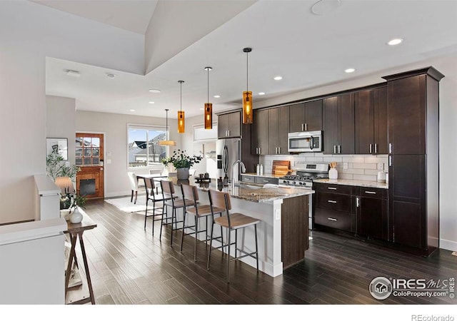 kitchen featuring light stone counters, hanging light fixtures, dark brown cabinets, appliances with stainless steel finishes, and an island with sink