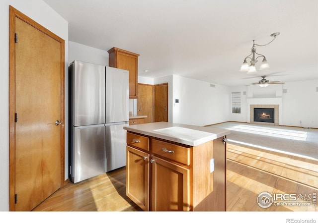 kitchen with stainless steel fridge, a fireplace, a kitchen island, ceiling fan with notable chandelier, and light wood-type flooring