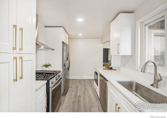 kitchen featuring appliances with stainless steel finishes, light wood-type flooring, wall chimney range hood, sink, and white cabinets