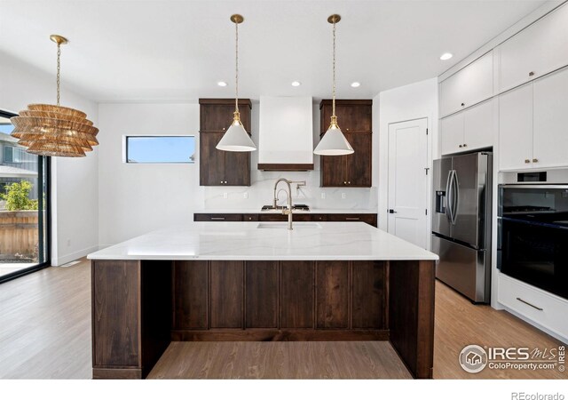 kitchen featuring stainless steel appliances, custom range hood, a kitchen island with sink, white cabinets, and dark brown cabinets