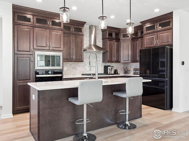 kitchen with dark brown cabinetry, built in microwave, stainless steel oven, wall chimney range hood, and a center island with sink