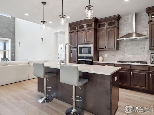 kitchen featuring dark brown cabinetry, stainless steel appliances, a kitchen island with sink, and wall chimney range hood