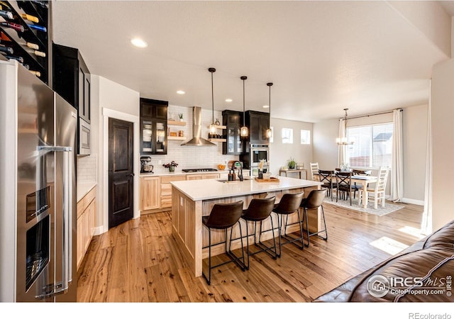 kitchen featuring sink, appliances with stainless steel finishes, an island with sink, wall chimney exhaust hood, and light brown cabinets