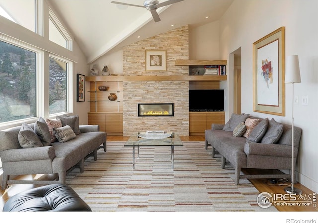 living area featuring ceiling fan, high vaulted ceiling, a stone fireplace, and wood finished floors