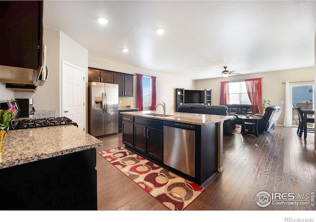 kitchen featuring stainless steel appliances, open floor plan, a sink, an island with sink, and light stone countertops