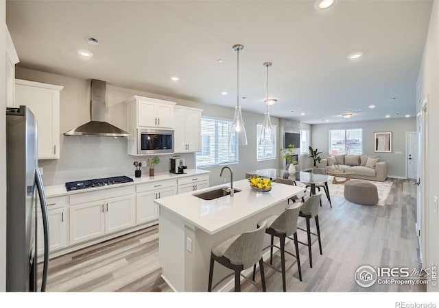 kitchen with stainless steel appliances, sink, wall chimney range hood, decorative light fixtures, and white cabinetry