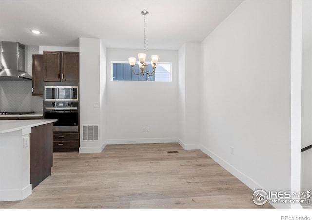 kitchen with wall chimney exhaust hood, decorative light fixtures, stainless steel appliances, tasteful backsplash, and light wood-type flooring