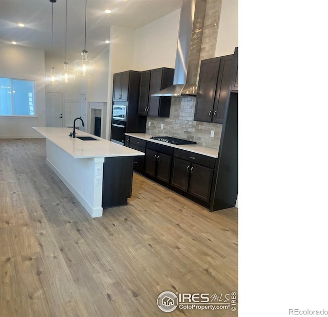 kitchen with wall chimney exhaust hood, sink, gas cooktop, an island with sink, and pendant lighting