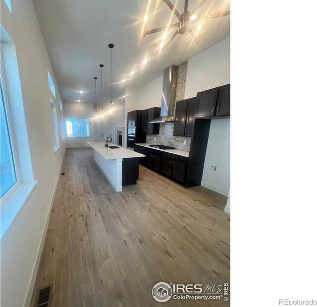 kitchen featuring wall chimney exhaust hood, sink, hanging light fixtures, an island with sink, and backsplash