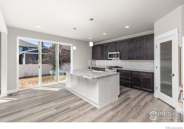 kitchen with an island with sink, hanging light fixtures, stainless steel appliances, light stone countertops, and light hardwood / wood-style flooring