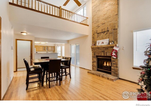 dining space with ceiling fan, light wood-type flooring, a towering ceiling, and a brick fireplace