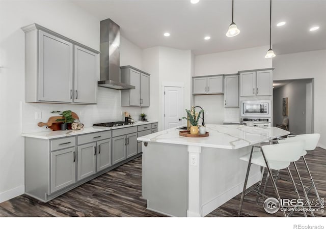 kitchen with gray cabinetry, wall chimney range hood, an island with sink, pendant lighting, and appliances with stainless steel finishes