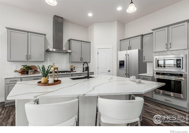 kitchen with sink, wall chimney exhaust hood, light stone counters, an island with sink, and appliances with stainless steel finishes