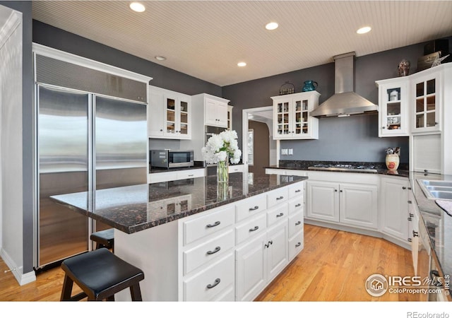 kitchen with appliances with stainless steel finishes, a kitchen island, wall chimney range hood, light hardwood / wood-style floors, and white cabinetry