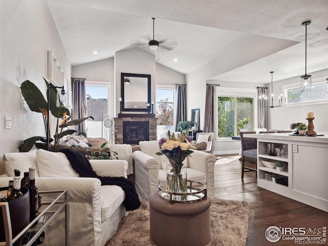 living room with dark wood-type flooring, vaulted ceiling, a textured ceiling, a fireplace, and ceiling fan with notable chandelier