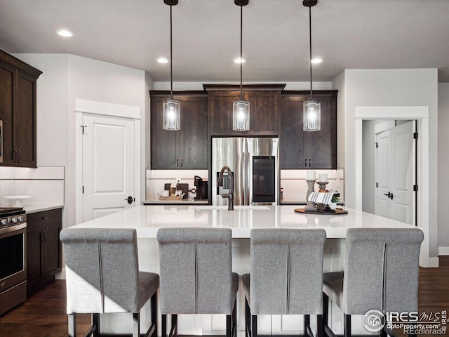 kitchen with dark brown cabinetry, backsplash, decorative light fixtures, a kitchen island with sink, and appliances with stainless steel finishes