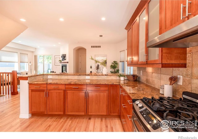 kitchen with stainless steel gas range oven, ventilation hood, decorative backsplash, light stone counters, and kitchen peninsula