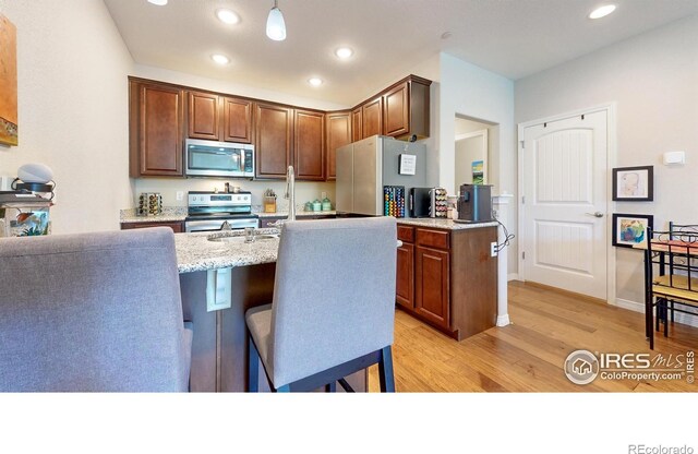 kitchen featuring light stone counters, an island with sink, pendant lighting, stainless steel appliances, and light hardwood / wood-style floors