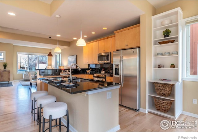 kitchen with light brown cabinets, a center island with sink, light hardwood / wood-style flooring, decorative light fixtures, and stainless steel appliances