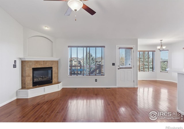 unfurnished living room featuring wood-type flooring, ceiling fan with notable chandelier, a healthy amount of sunlight, and a tiled fireplace